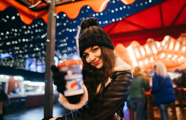 Pretty positive girl in warm clothes stands in the evening under an umbrella at the fair with a hot drink in her hand, offers it to camera and smiles.Cute lady shows a warming drink to the camera.