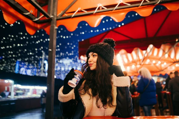 夜に傘の下に光で飾られた冬のフェアの女性は 熱い飲み物を飲み ガーランドによって照らされた夜の通りの背景を離れて見ます クリスマスマーケット — ストック写真