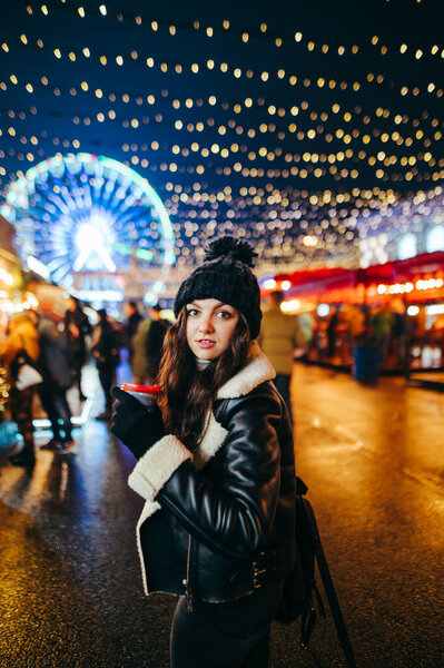 Pretty girl in warm clothes walks on the evening decorated with lanterns fair on the background of the street market and ferris wheel. Lady at the Christmas market with a warming drink in hand.