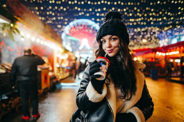 美しい正の女性は フェアで魅力的なクリスマスストリートに立って 暖かい飲み物を飲み 彼女の顔に笑顔でカメラを見て 暖かい冬の服を着ています 新年とクリスマス — ストック写真