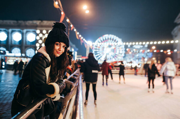 Positive girl in a hooded dress and hat stands in the evening near the rink on the background of a decorated street and a Ferris wheel, looks into the camera and smiles. Walk at Christmas.