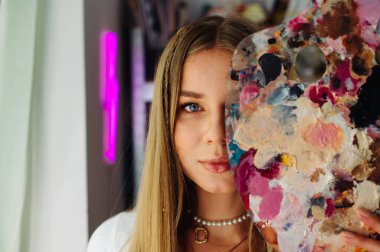 Ressamın yaratıcı yakın plan portresi, yağlı boyayı karıştırmak için yüzünü paletle kaplıyor ve kameraya bakıyor. Kadın sanatçı yüzünün yarısını paletle kaplamış..