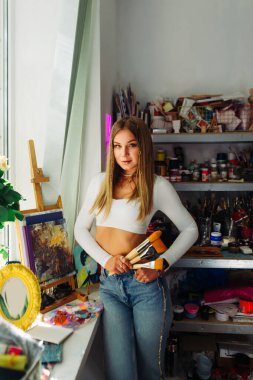 Atölyede elinde fırçalarla yaratıcı kadın ressamın portresi, arkaplandaki raflara resim aletleriyle bakıyor..