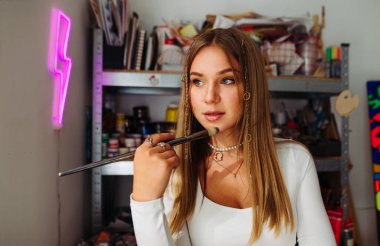 Atölyedeki kameraya poz veren çekici kadın sanatçı, elinde fırçayla arka planda resim yapmak için gerekli aletlerle.