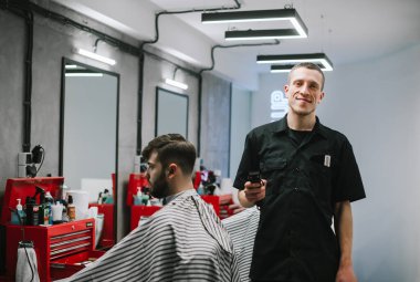 İş yerinin yanında duran gülümseyen berberin portresi ve elinde kırpıcı ile kameraya bakıp gülümseyen bir müşteri. İş yerinde kameraya poz veren pozitif erkek kuaför. Kuaförlük konsepti.