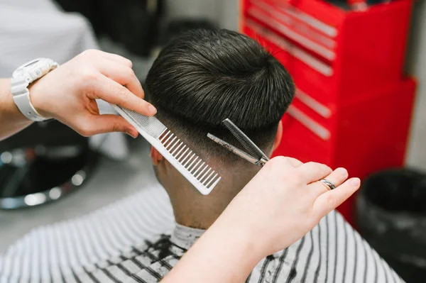 Hands of a professional barber with scissors and comb combs the hair of a brunette man, close photo. Process of creating a fashionable haircut for a client at a men's hairdresser.