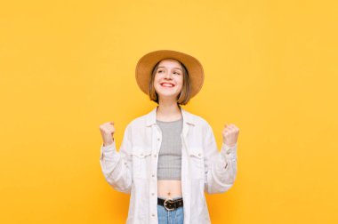 Güneş şapkalı ve yazlık elbiseli neşeli kadın portresi sarı bir arka planda izole edilmiş, sevinç ve gülümseme görmek için ellerini kaldırmış. Turist kız turuncu arka planda duruyor. Boşluğu kopyala