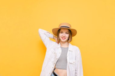Beyaz gömlekli, güneş şapkalı, sarı arka planda duran oldukça olumlu bir kız yüzünde bir gülümsemeyle kameraya bakıyor. Sarı arka planda poz veren yaz kıyafetleri içindeki yürüyüşçü kız. İzole edilmiş.