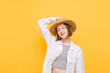 Şapkalı ve açık renk elbiseli gülümseyen kadının portresi sarı arka planda duruyor. Gözleri kapalı ve mutlu yüzlerden hoşlanıyor, şapkaya dokunuyor. İzole edilmiş. Boşluğu kopyala