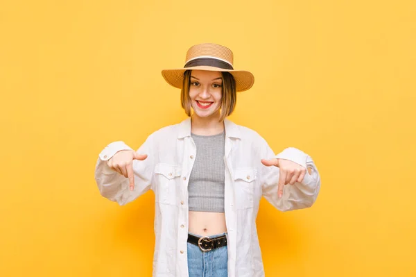 Happy tourist girl in light clothing and hat, looks into the camera with smile on face and points fingers down at copy space. Smiling lady in summer clothes showing fingers down on yellow background.