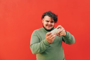 Konsantre olmuş, neşeli İspanyol adam akıllı telefonunu tutuyor ve kendini eğlendirmek için dijital teknolojiler kullanarak cihazda oyun oynuyor. Kırmızı bir arkaplanda çekilmiş..