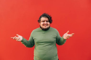 Şaşkın, genç, İspanyol sakallı, kilolu adam ellerini kaldırarak, kaşlarını çatarak, şaşkın ve kararsız hissederek şaşkınlığını ifade ediyor. Kırmızı arkaplan.