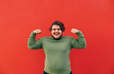 Güçlü ve komik genç şişman İspanyol adam ellerini kaldırarak ve yumruklarını sıkarak, kaslarını göstererek, gülümseyerek ve kırmızı duvarın önünde durarak gücünü gösteriyor..