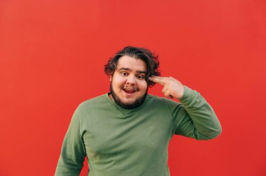 Komik, şişman, sakallı genç adam şaka gibi bir şekilde elinde silah taklit ediyor, deli ve garip görünüyor, kırmızı duvarın önünde duruyor..