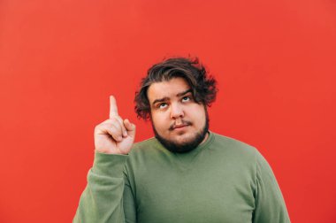 Düşünceli, şişman, sakallı İspanyol bir adamın portresi. Parmağını kaldırarak, yukarı bakarak, merak ederek, yaratıcılığını kullanarak, kırmızı arka planda durarak bir fikir jesti yapıyor..