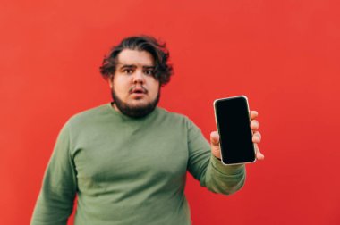 Şaşırmış ve şaşırmış genç aşırı kilolu adam elinde cihazla izleyiciye siyah ekranlı bir akıllı telefon gösteriyor. Telefona odaklan..