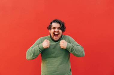 Neşeli ve mutlu sakallı genç adam başarısını ve başarısını kutluyor, kazandıktan sonra sevinçle bağırıyor, kırmızı arka planda duruyor..