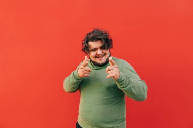 Neşeli, çekici İspanyol genç adam kırmızı duvarın önünde duruyor ve parmaklarını kameraya ve izleyicilere doğrultuyor. Olumlu ve arkadaş canlısı..