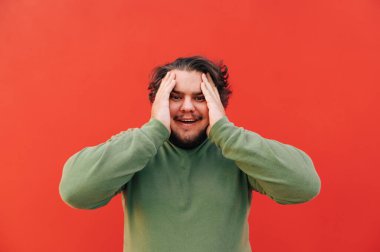 Mutlu İspanyol genç adam duygusal bir rahatlama ve heyecan hissediyor, kırmızı arka planda duruyor, kafasına dokunuyor, neşeli oluyor..