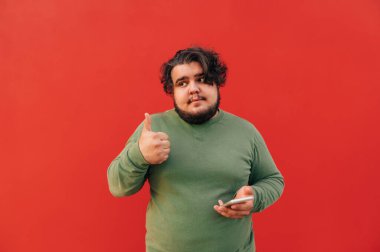 Sevimli ve çekici sakallı, kilolu, kırmızı arka planda, akıllı telefonunu kaldırmış, yan bakıyor..