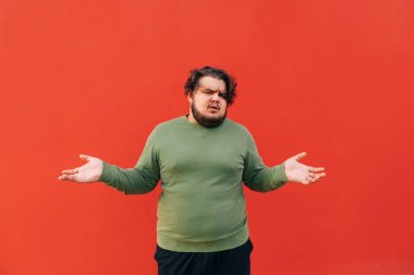 Kafası karışmış, sakallı, İspanyol bir adam ellerini kaldırıyor umutsuzluk içinde, rahatsız ve şaşkın hissediyor, somurtuyor, izole bir kırmızı arka planda duruyor..