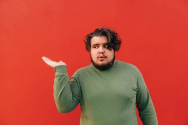 Komik ve eğlenceli genç İspanyol adam elinde kırmızı bir arka planla bir ürünün reklamını yapıyor, izleyicinin ilgisini çekmeye çalışıyor..