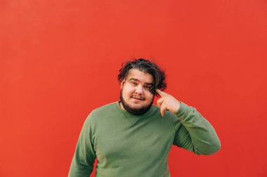 Neşeli obez İspanyol genç adam parmağını kafasına doğrultuyor, düşünceli bir hareket sergiliyor, odaklanıyor ve konsantre oluyor, kırmızı arka planda duruyor..