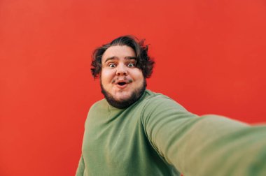 Heyecanlı ve neşeli İspanyol bir adam kameraya selfie çekiyor, ağzı açık, kırmızı bir arka planda dururken şaşırmış bakıyor..