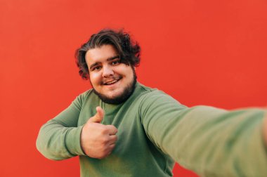 Neşeli, karizmatik genç adam kameranın önünde parmağını kaldırmış, mutlu, gülümseyen ve kırmızı duvarın önünde duran bir selfie çekiyor..