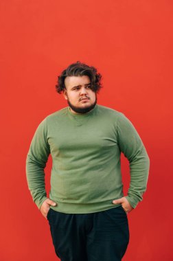 Şık saç kesimi ve sakallı yakışıklı bir adamın dikey çekimi. İzole edilmiş kırmızı bir arka planda duruyor. Elleri cebinde, yana bakıyor..