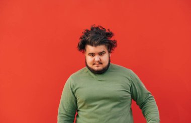 Kıvırcık saçlı komik şişman sakallı adam kırmızı duvarın önünde duruyor, yeşil kazak giyiyor, mutlu ve komik..