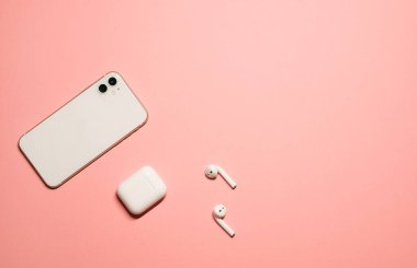Pastel pembe arka planda, beyaz bir akıllı telefon ve şarj çantalı kablosuz kulaklıklı düz bir fotoğraf..