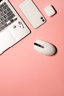 Üzerinde gümüş gri dizüstü bilgisayarı, modern akıllı telefonu, beyaz plastik faresi ve pembe arka planda şarj çantası olan kablosuz kulaklığı olan dikey düz bir fotoğraf vardı..