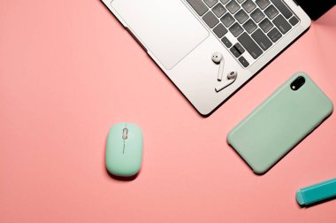 Turkuaz bir bilgisayar faresi, açık mavi silikon çantalı akıllı telefon, gümüş dizüstü bilgisayar, kablosuz kulaklık ve pembe arka planda mavi işaret..