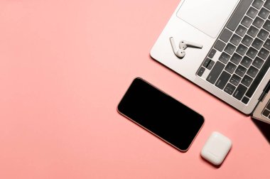 Gri dizüstü bilgisayarı, dokunmatik ekranlı cep telefonu ve izole edilmiş pembe arka planda şarj çantası olan kablosuz kulaklığı olan modern teknoloji dairesi..