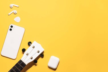 Beyaz ukulele fretboard, modern cep telefonu, şarj kutusu olan kulaklıklar ve gitar penalı sarı arka planda bir kopya alanı olan düz bir kompozisyon..