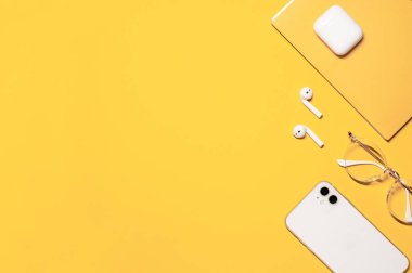 Solunda fotokopi alanı olan düz bir fotoğraf ve sağında nesneler: beyaz akıllı telefon, şarj kutusu olan kablosuz kulaklık, şık gözlükler ve sarı arka planda sarı bir defter..
