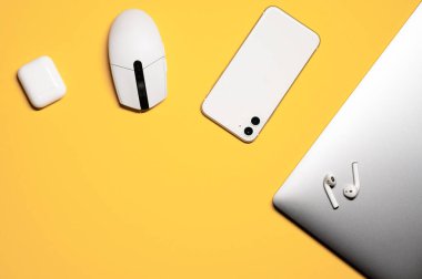 Dijital cihazlarla basit düz konum: beyaz akıllı telefon, şarj çantalı kablosuz kulaklık, beyaz renkli plastik fare ve sarı arkaplanda gümüş bir laptop..