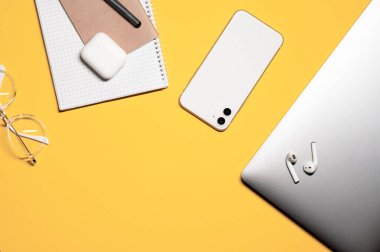 Sarı arka planda gümüş dizüstü bilgisayarı, beyaz akıllı telefonu, defterleri, kalemi, şık gözlükleri ve kablosuz kulaklıkları olan renkli bir kompozisyon..