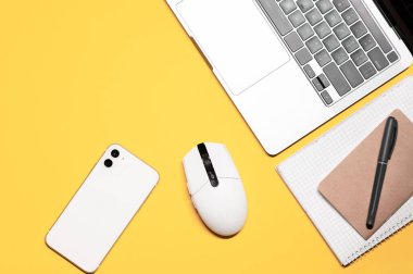 Beyaz kablosuz bilgisayar faresi, gümüş gri laptop, beyaz akıllı telefon, iki not defteri ve sarı arka planda bir kalem..