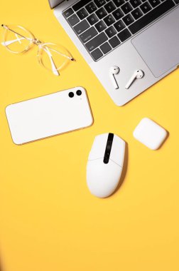Beyaz bilgisayar faresi, siyah klavyeli modern gri dizüstü bilgisayar, beyaz akıllı telefon, gözlük ve renkli sarı arka planda kablosuz kulaklık..
