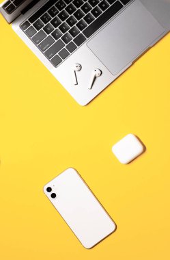 Gri dizüstü bilgisayarı, beyaz cep telefonu ve şarj çantalı kablosuz kulaklığı olan sarı arka planda dikey düz bir kompozisyon..