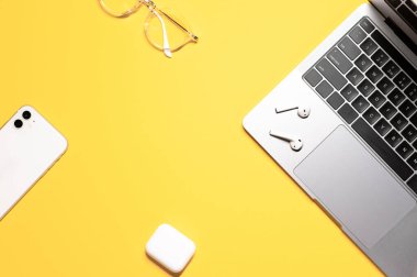 Gri dizüstü bilgisayarı, beyaz cep telefonu, gözlüğü ve kablosuz kulaklığı olan sarı arka planda güzel düz bir kompozisyon..