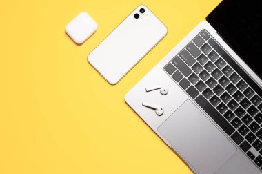 Gümüş gri dizüstü bilgisayarı, beyaz cep telefonu, kablosuz kulaklığı ve sarı arka planda şarj çantası olan düz bir kompozisyon..