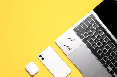 Tasarım, modern gri dizüstü bilgisayarı olan işyeri taklidi, şarj kutusu olan kulaklıklar ve sarı arka planda beyaz bir akıllı telefon..