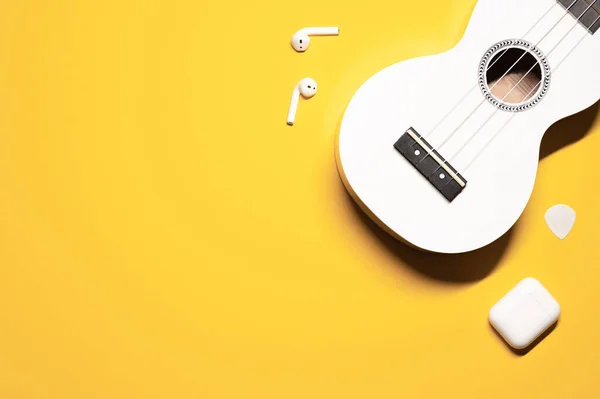 Simple layout composition on a colorful yellow background with a musical instrument - ukulele soprano with nylon strings, wireless earphones with a charging case and a guitar pick.