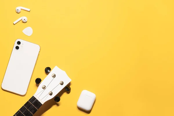 Flat lay composition with a copy space on a yellow background with a white ukulele fretboard, modern mobile phone, earphones with a charging case, and a guitar pick.