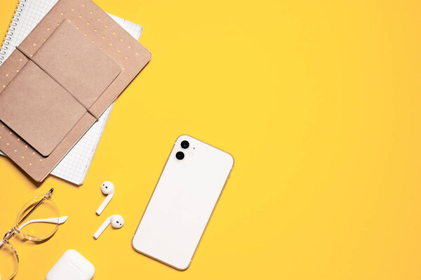 Colorful flat lay shot with a stack of notepads and diaries, modern smartphone, stylish eyeglasses, and wireless earphones with a charging case on a yellow background.
