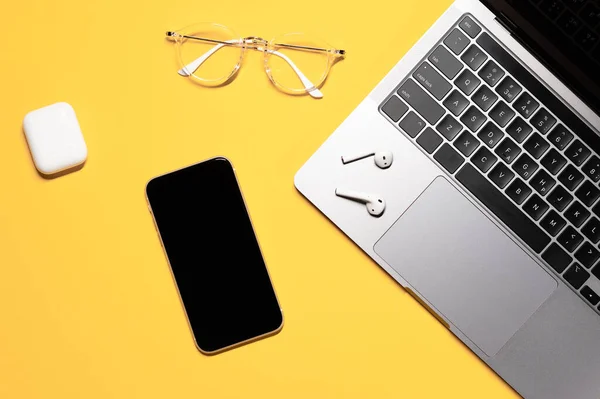 Creative layout with a smartphone with touch screen, silver grey laptop with a black keyboard, stylish goggles, and wireless earphones on a yellow background.