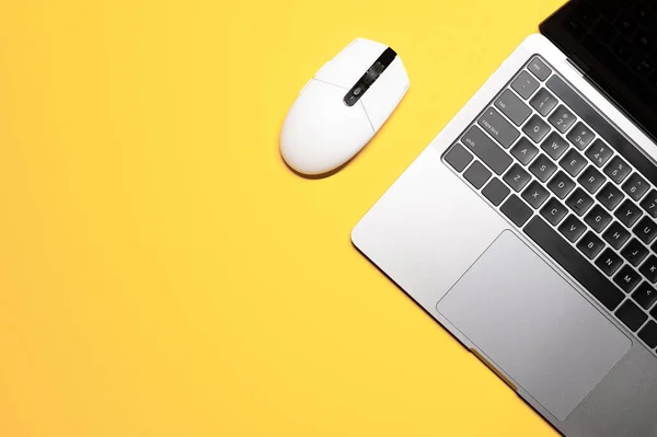 Minimalist layout shot with a simple composition: a silver grey laptop computer with a black keyboard, and a plastic white mouse on a yellow background.